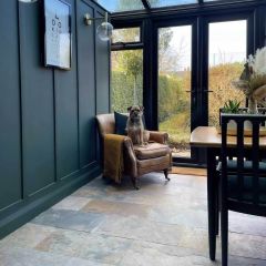 Conservatory space with light filled windows and roof, brown leather chair, dog, and rustic stone floor tiles in natural warm tones.