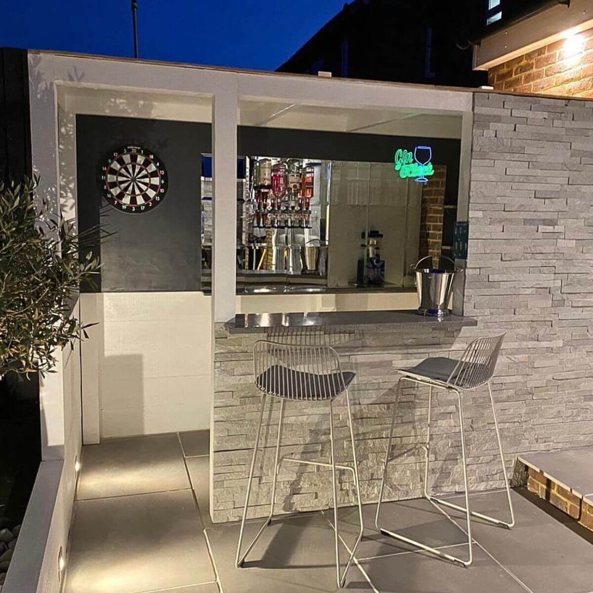 Outdoor bar clad in ivory split face tiles, two bar stools, coctail bar and dart board at night